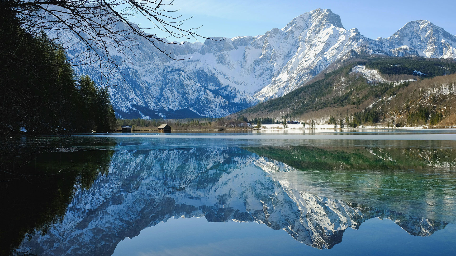 Snowy mountains are reflected in a lake with wooden huts on the shore. 