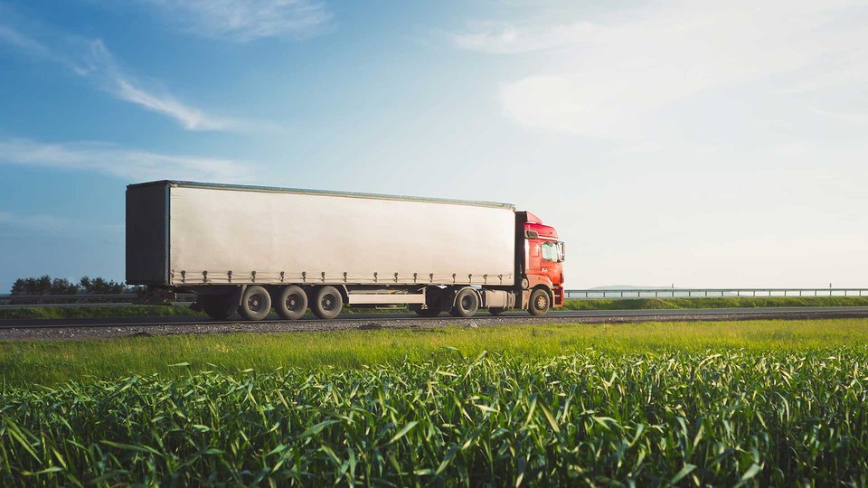 A red truck with a trailer is driving next to a green field on a country road.