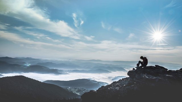 Person sitting on a rock with a view of a misty mountain landscape under a bright sun.
