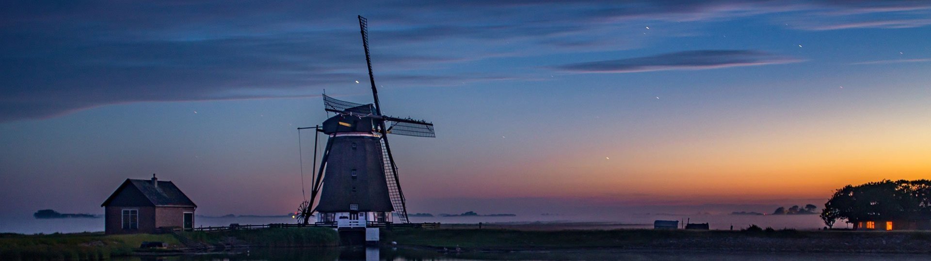 A windmill and a small hut by the water at sunset with a reflection in the lake.