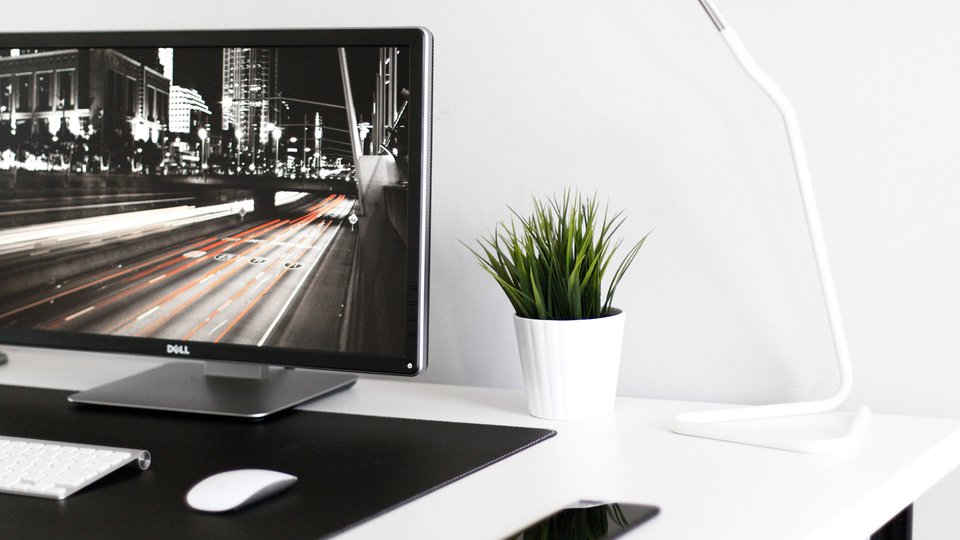 A minimalist desk with a monitor, with a plant and a white lamp on a white desk.