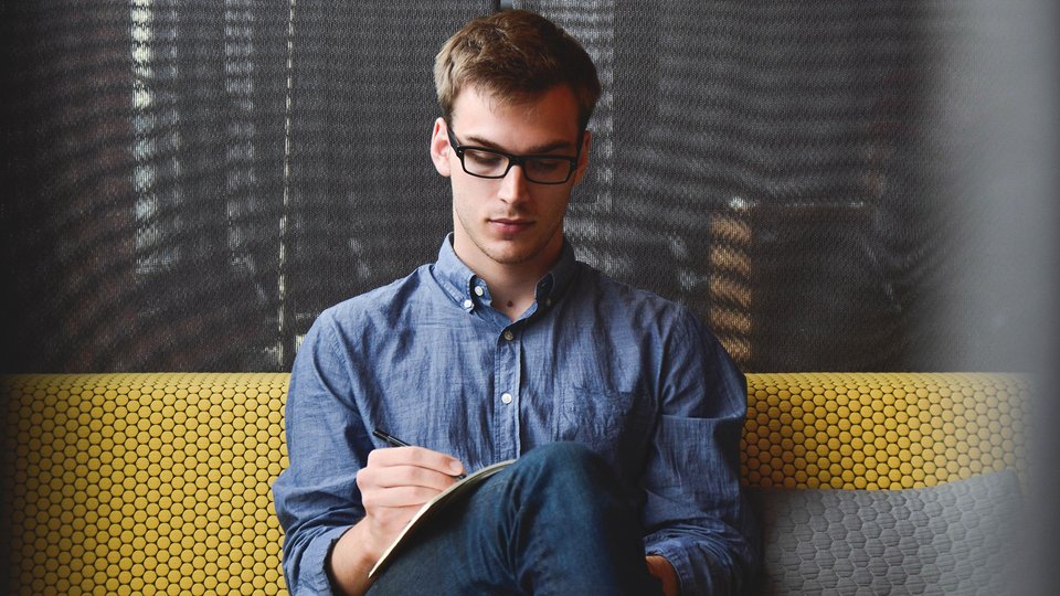 Young man with glasses writing intently in a notebook on a yellow upholstered bench.