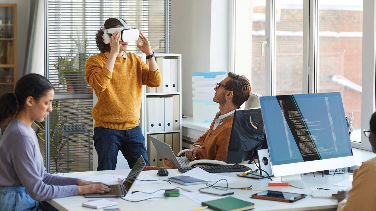 Employees in an office test VR glasses and work on computers and laptops.