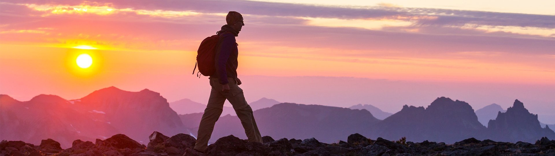 Person hiking at sunset on a rocky mountain landscape with mountain peaks in the background.
