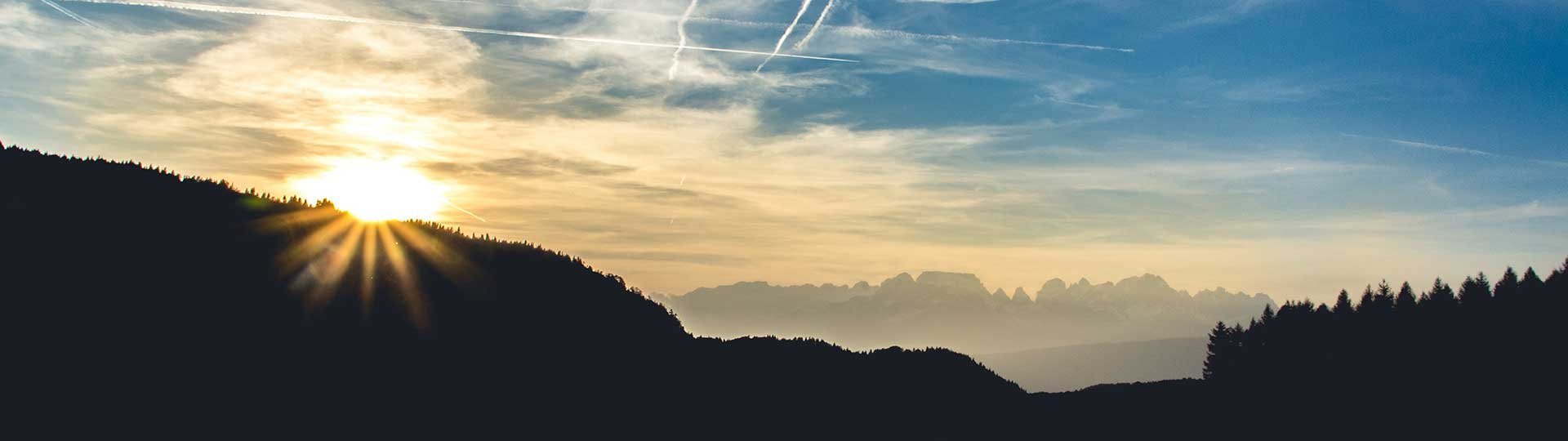 Sunrise behind a wooded hill with vapor trails in the sky and a mountain panorama.