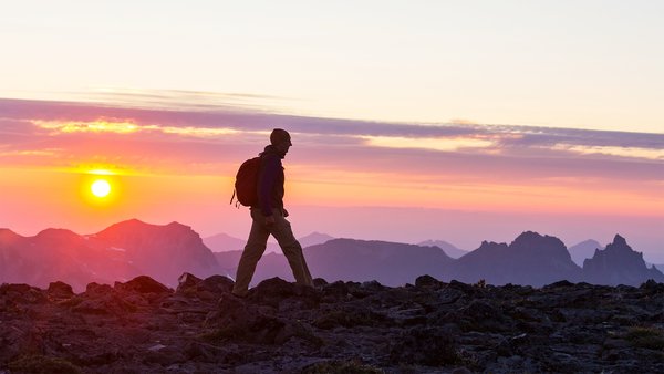 Person hiking at sunset on a rocky mountain landscape with mountain peaks in the background.