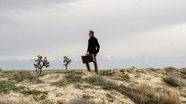 Man with briefcase stands in desert landscape with mountains in the background.