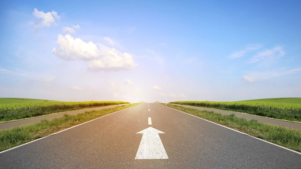 A straight road with a white arrow leads into the horizon, surrounded by green fields.