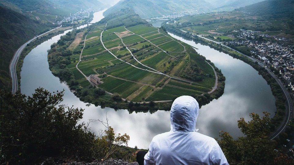 Person in a white jacket looks out over a picturesque bend in the river with vineyards.