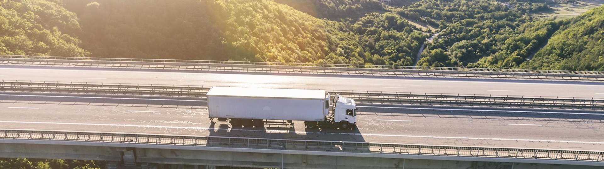 White truck drives over a bridge in a mountainous landscape in sunlight.