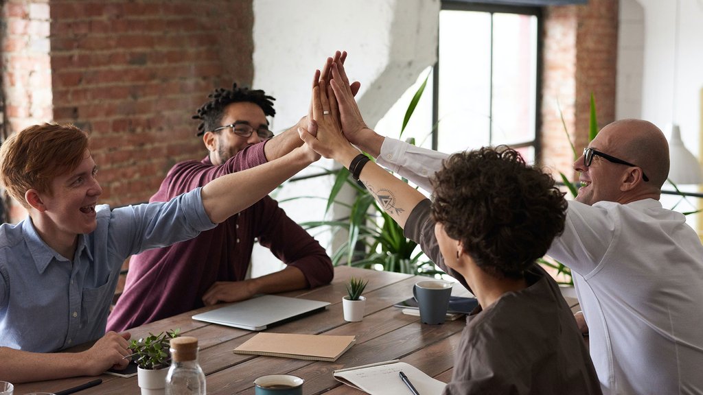 Four team members laugh and give each other high-fives during a meeting at a wooden table.