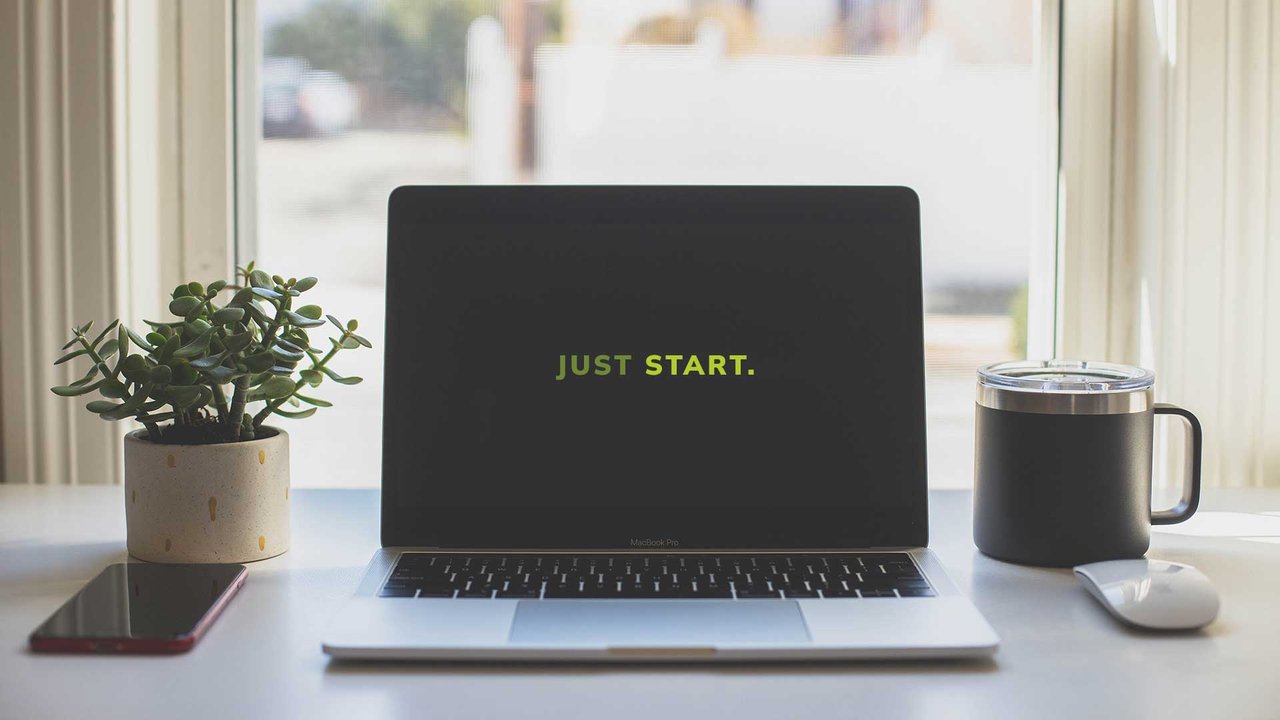 Minimalist desk with a MacBook which says “JUST START.” on the screen an a mug next to it. 
