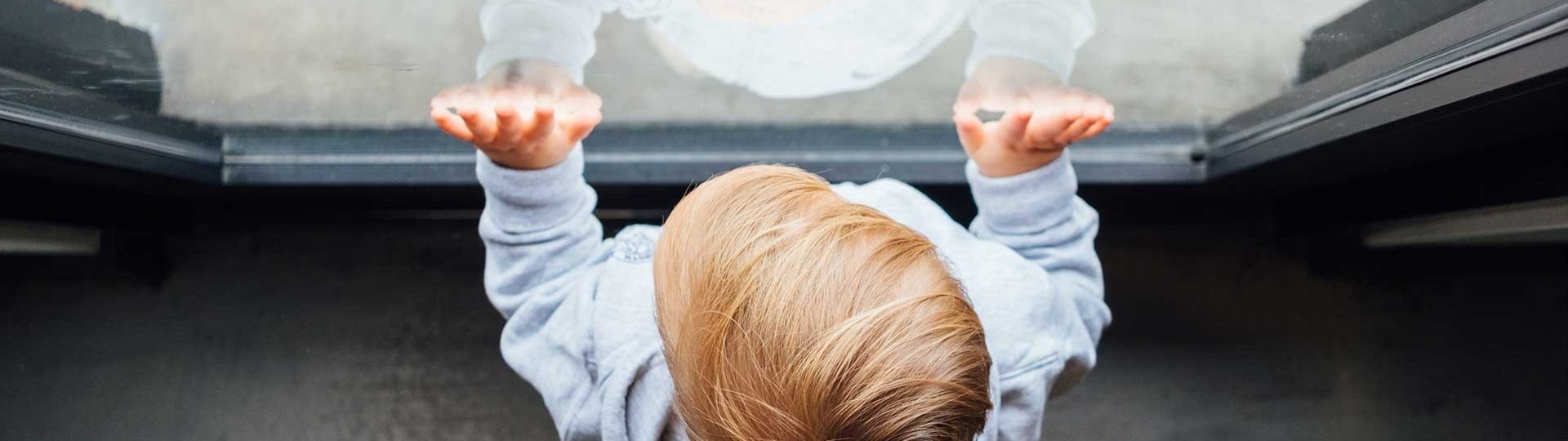 Small child touches a window and looks outside, reflected in the glass.