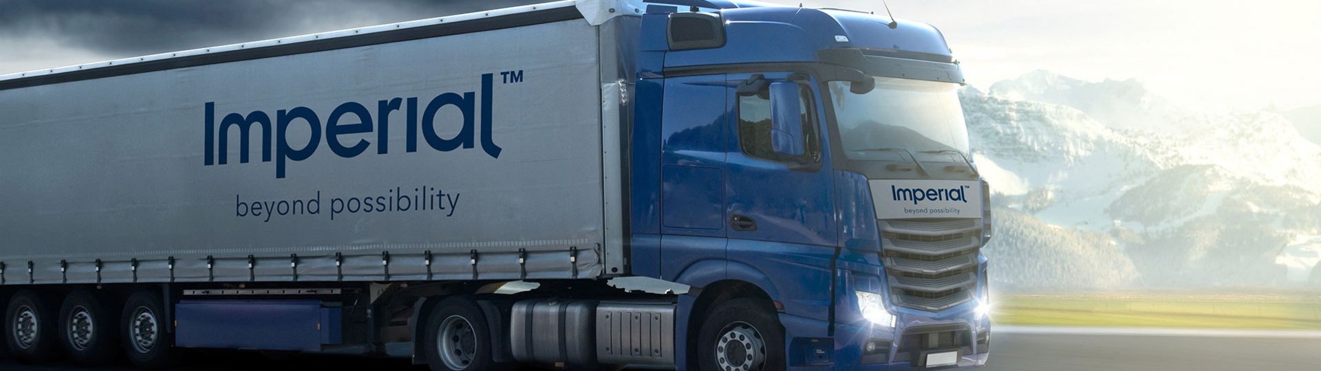 Blue Imperial truck driving on an open road under a cloudy sky against a mountain backdrop.