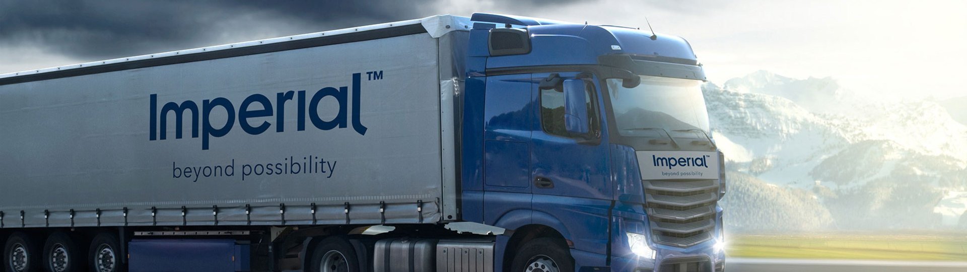 Blue Imperial truck driving on an open road under a cloudy sky against a mountain backdrop.