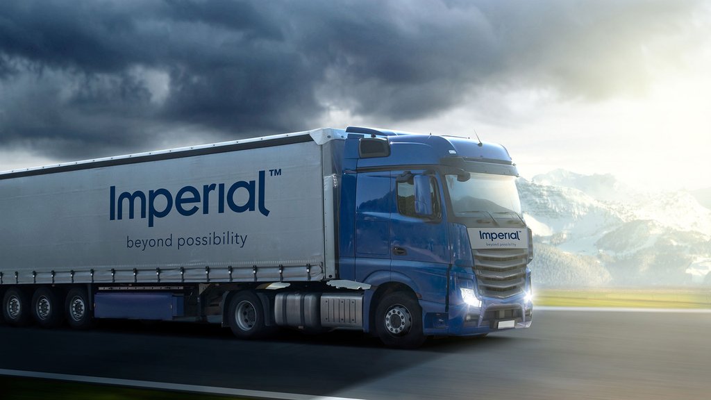 Blue Imperial truck driving on an open road under a cloudy sky against a mountain backdrop.