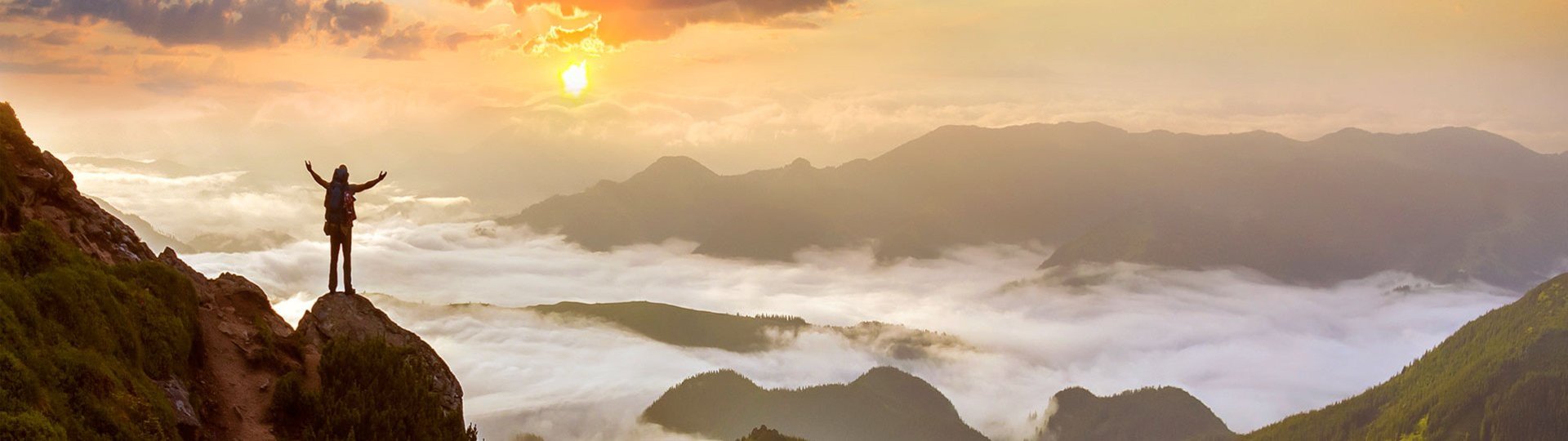 A person stands on a mountain peak with outstretched arms and gazes into a misty sunset.