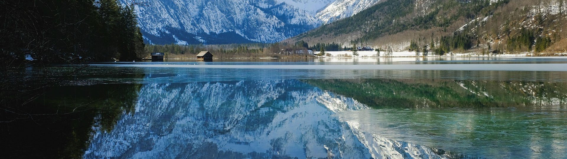 Snowy mountains are reflected in a lake with wooden huts on the shore.