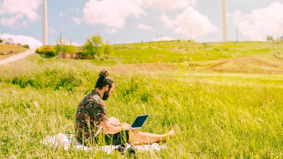 Person working with a laptop on a meadow, wind turbines in the background, shows sustainable work.