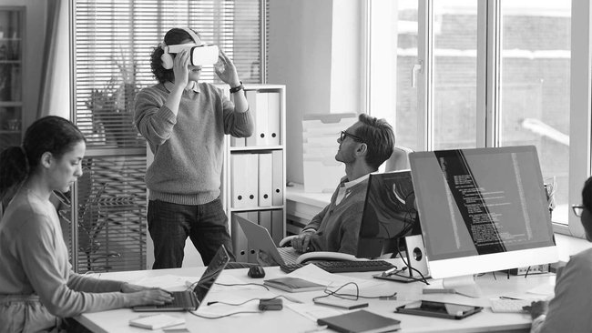 Employees in an office test VR glasses and work on computers and laptops.