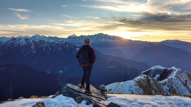Mountaineer with rucksack stands on summit and looks out over snow-covered mountains.
