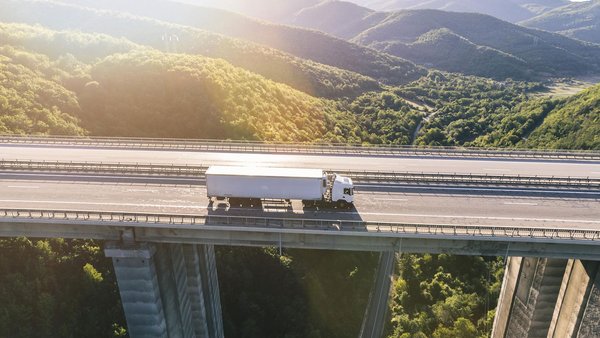 White truck drives over a bridge in a mountainous landscape in sunlight.