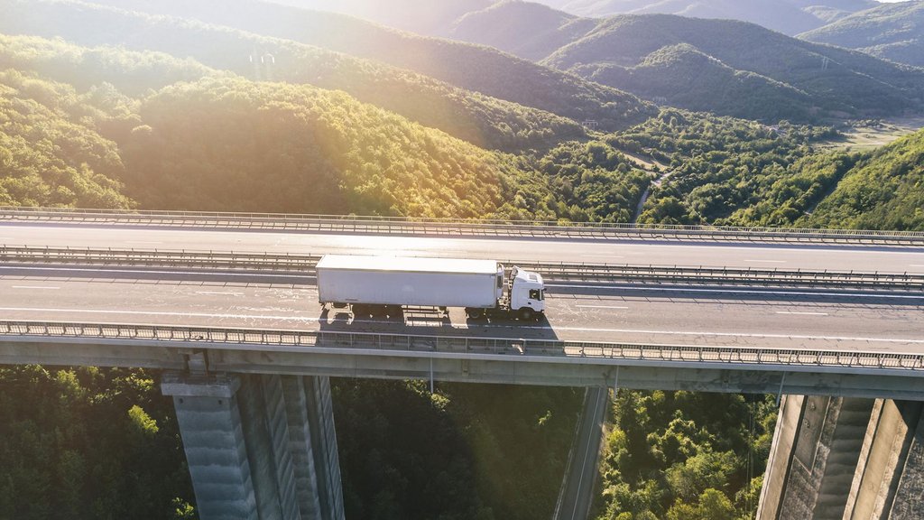 White truck drives over a bridge in a mountainous landscape in sunlight.