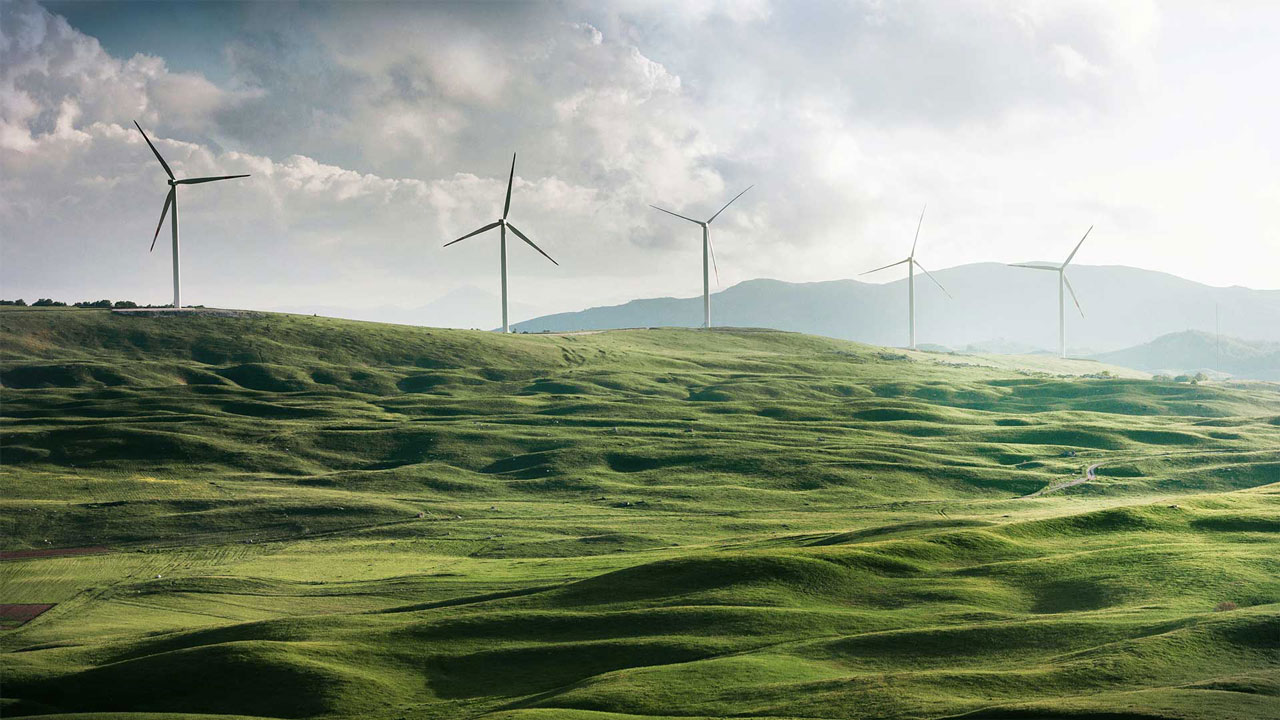 Wind turbines on green hills against a mountain backdrop under a cloudy sky.